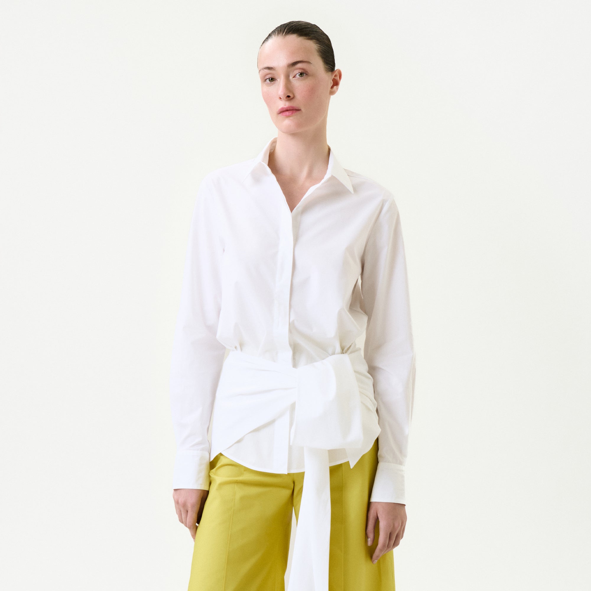 A person with slicked-back hair and a neutral expression stands against a plain white background, wearing the Malen Shirt—a white 100% cotton button-up with a large tied sash at the waist—paired with yellow wide-leg pants.