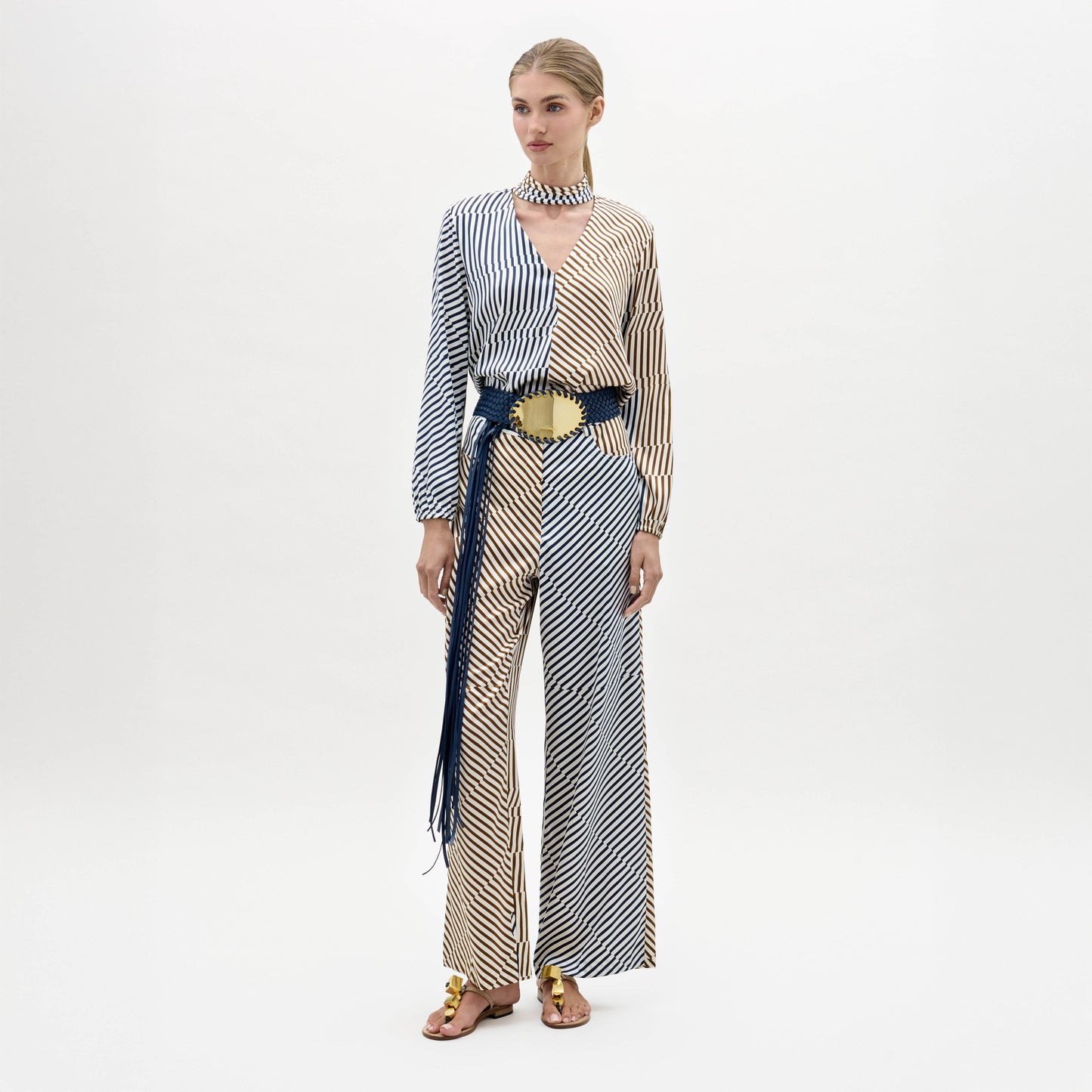 A woman stands against a plain background wearing the Emine Pant, styled with gold sandals and a matching choker.