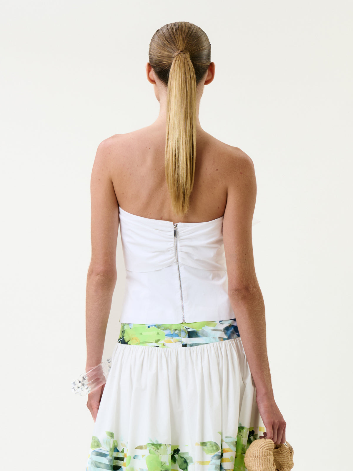 A woman with straight, long blond hair in a ponytail wears the Kacy Top—a white strapless top featuring a back zipper—paired with a white skirt adorned with green floral patterns, standing against a plain background.
