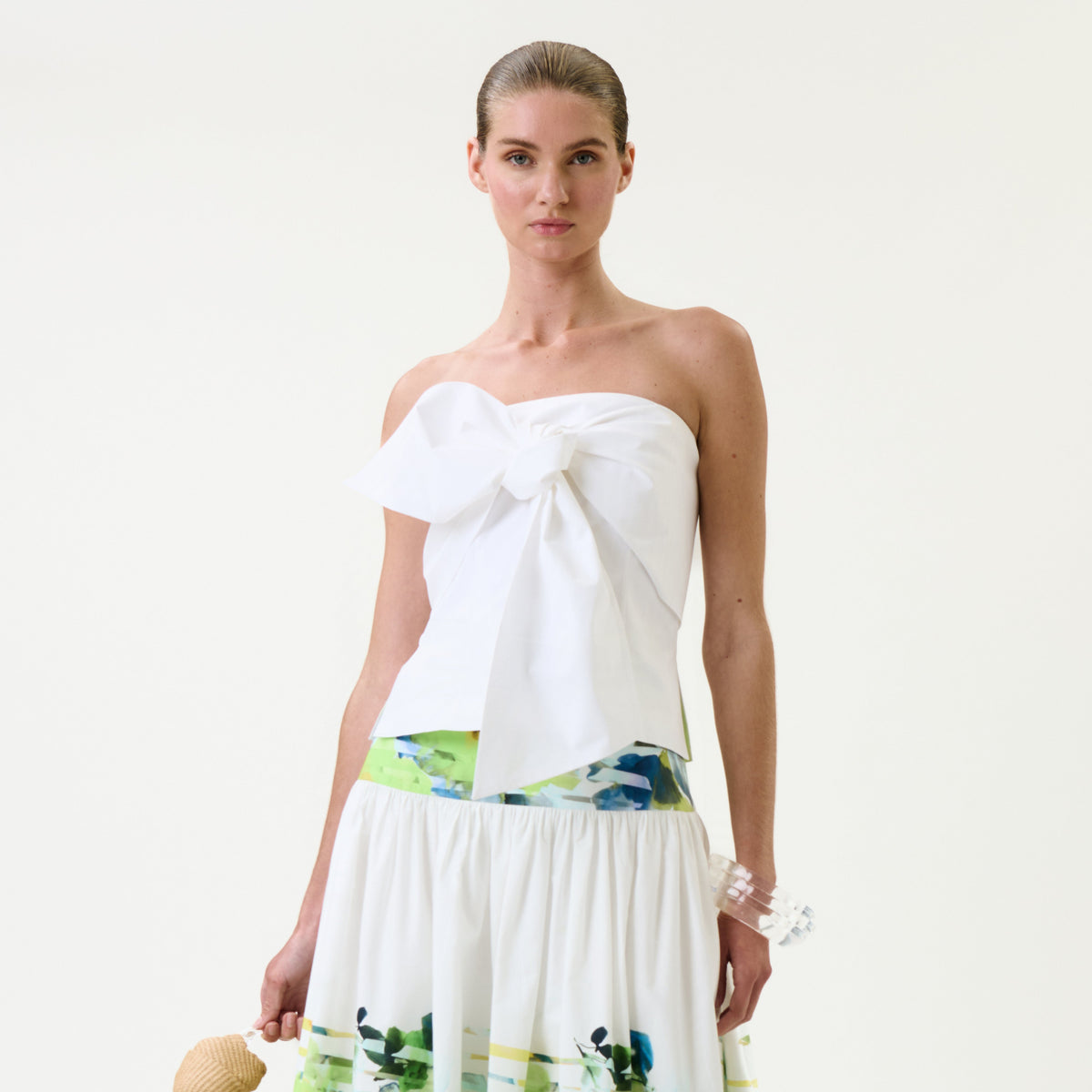 A woman wearing the Kacy Top, a strapless white blouse adorned with a large bow, and a white skirt featuring colorful prints stands against a plain background, holding a small straw bag.