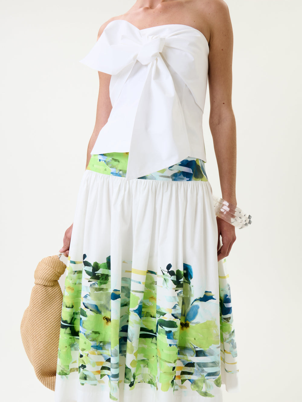 Woman wearing the Kacy Top, a strapless white top with a large bow, paired with a white skirt featuring green and blue abstract patterns, holding a tan textured bag and wearing a clear chunky bracelet.