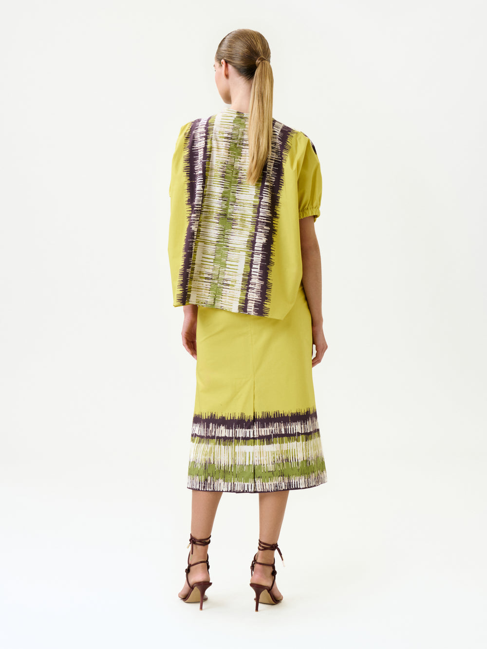 A woman with a low ponytail stands facing away, wearing the Atira Skirt—a yellow 100% cotton skirt and top set with abstract green, white, and purple stripes, a detachable belt—and brown strappy high-heeled sandals.