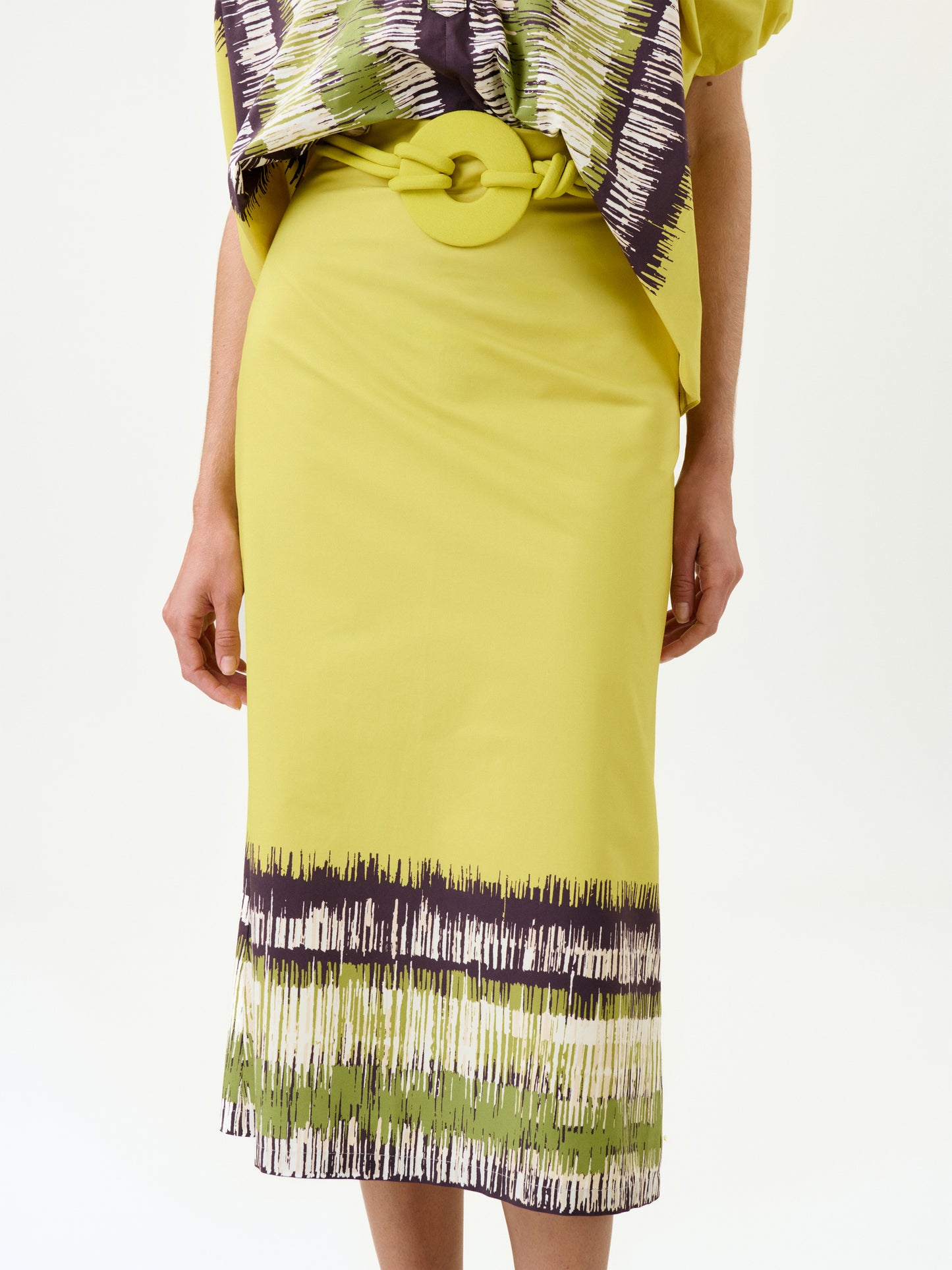 A person wears the Atira Skirt—yellow Italian fabric with an abstract green and brown pattern at the hem, a wide detachable belt, and a matching top. Cropped from shoulders down.