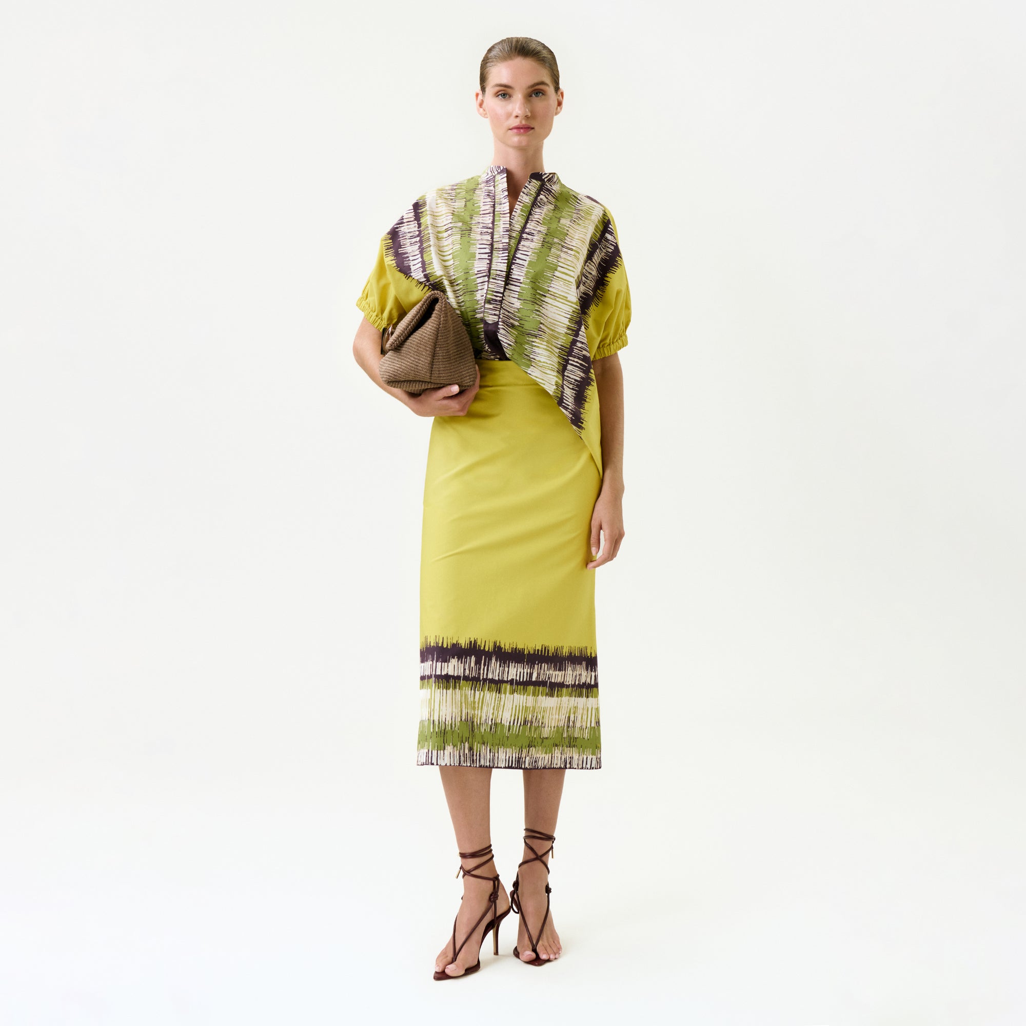 A woman stands against a plain background wearing the Atira Skirt, featuring abstract purple, white, and green patterns on yellow 100% cotton fabric, paired with brown strappy sandals and a large brown clutch under her arm.