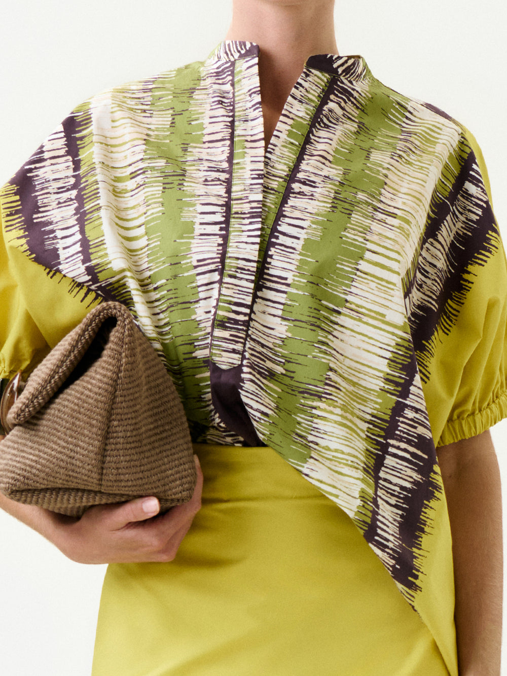 A person wears the Susane Blouse, featuring a green, white, and black abstract pattern made from Italian fabric, paired with a yellow dress and holding a brown woven clutch against a plain background.