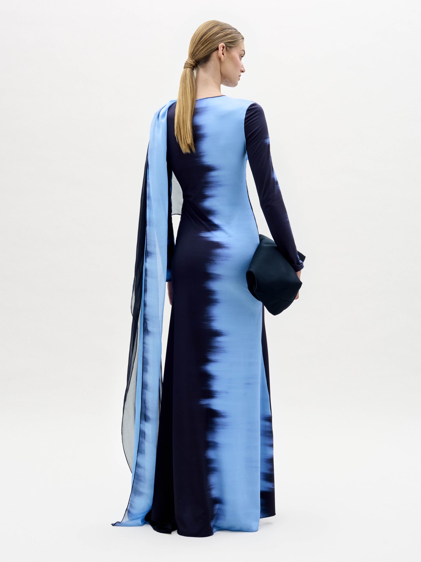 A person stands against a plain white background, wearing the Moon Dress—a long blue and black gradient gown with an attached flowing scarf—and holding a dark clutch.