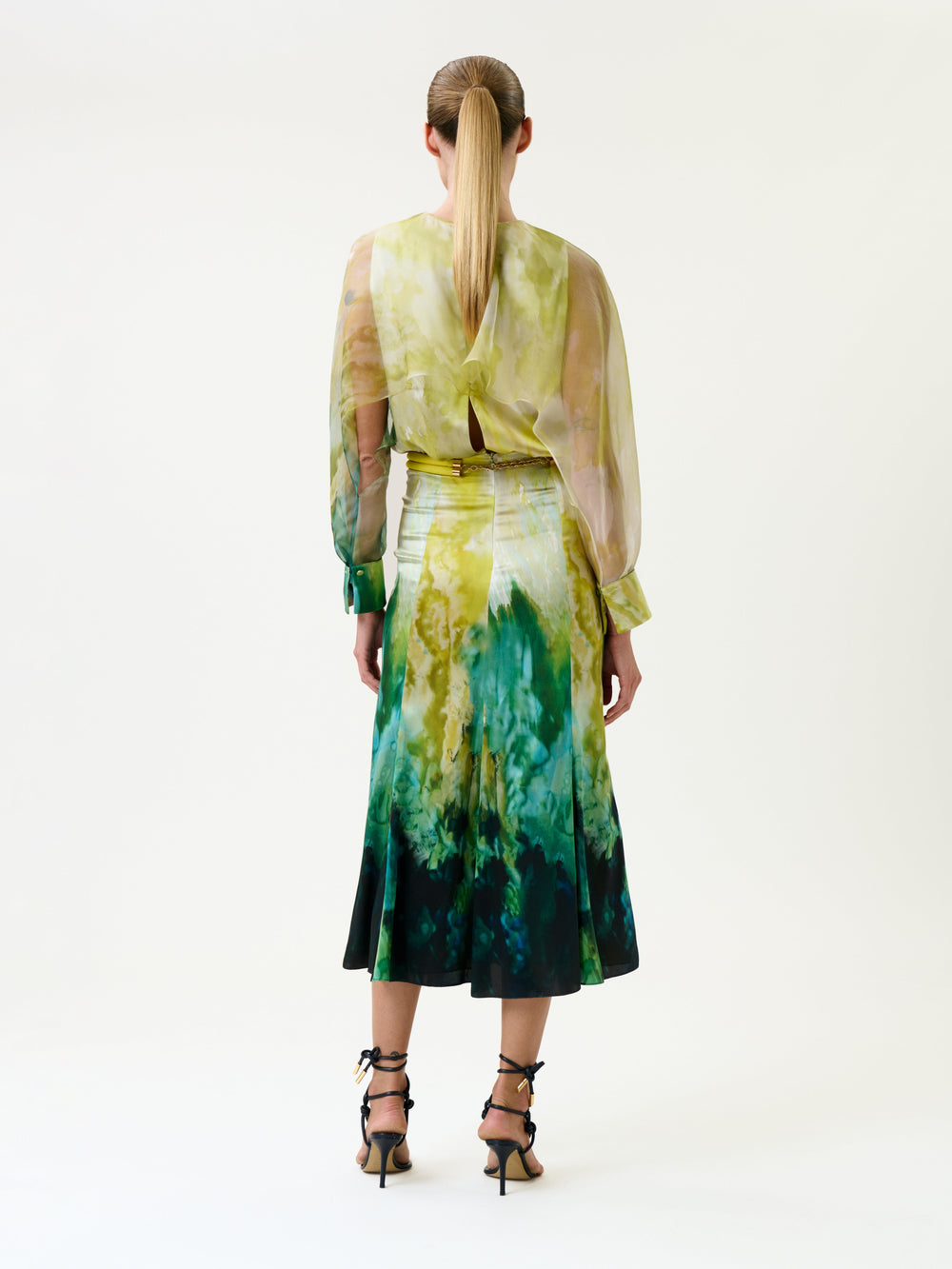 A woman with a ponytail stands facing away, wearing the Rubina Dress—a sheer long-sleeve blouse and a green-yellow tie-dye skirt—paired with black strappy heels against a plain white background.