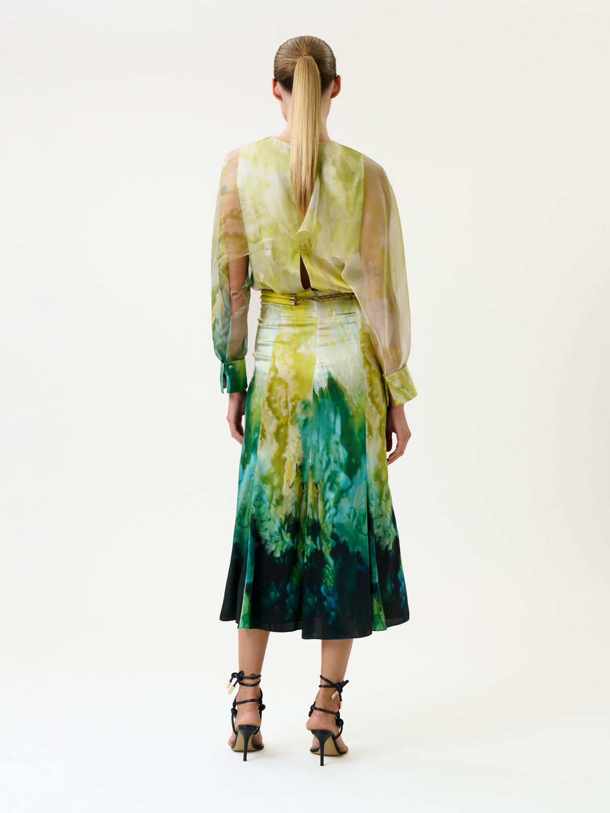A woman with a ponytail stands facing away, wearing the Rubina Dress—a sheer long-sleeve blouse and a green-yellow tie-dye skirt—paired with black strappy heels against a plain white background.