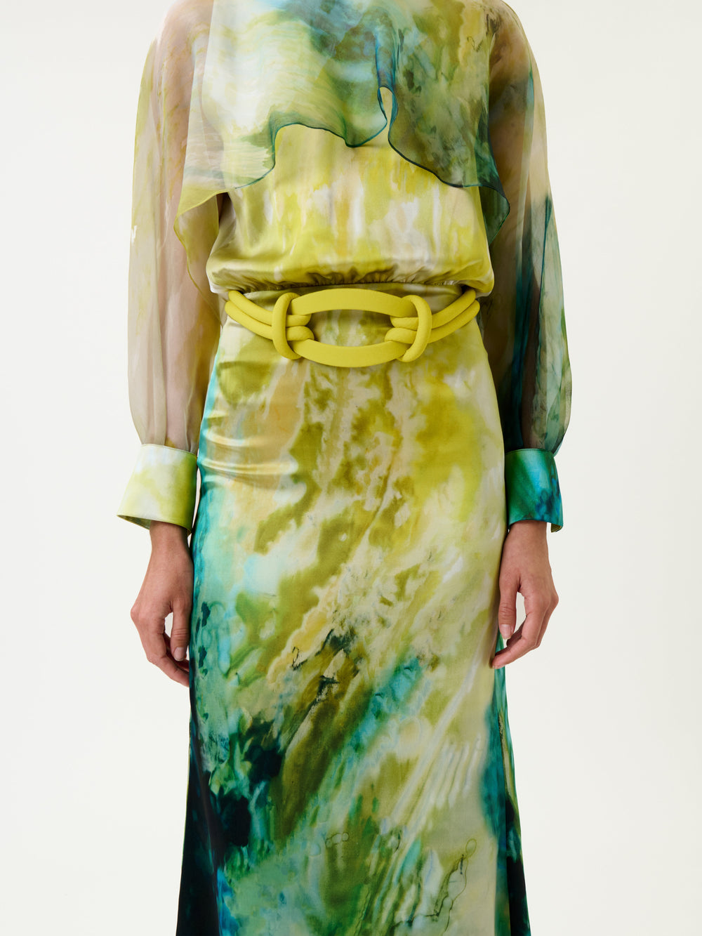 A woman wears the Rubina Dress, a long-sleeve design with a green, yellow, and blue abstract pattern and a matching wide belt featuring a large oval buckle. Only her torso, arms, and upper legs are visible against a plain background.