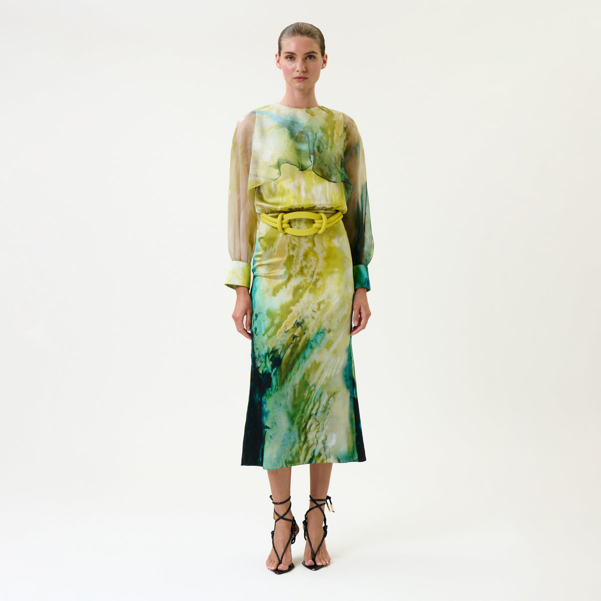 A woman wears the Rubina Dress, a long-sleeved, watercolor-patterned piece in green, yellow, and blue tones, belted at the waist. She pairs it with strappy black heels and stands against a plain white background.