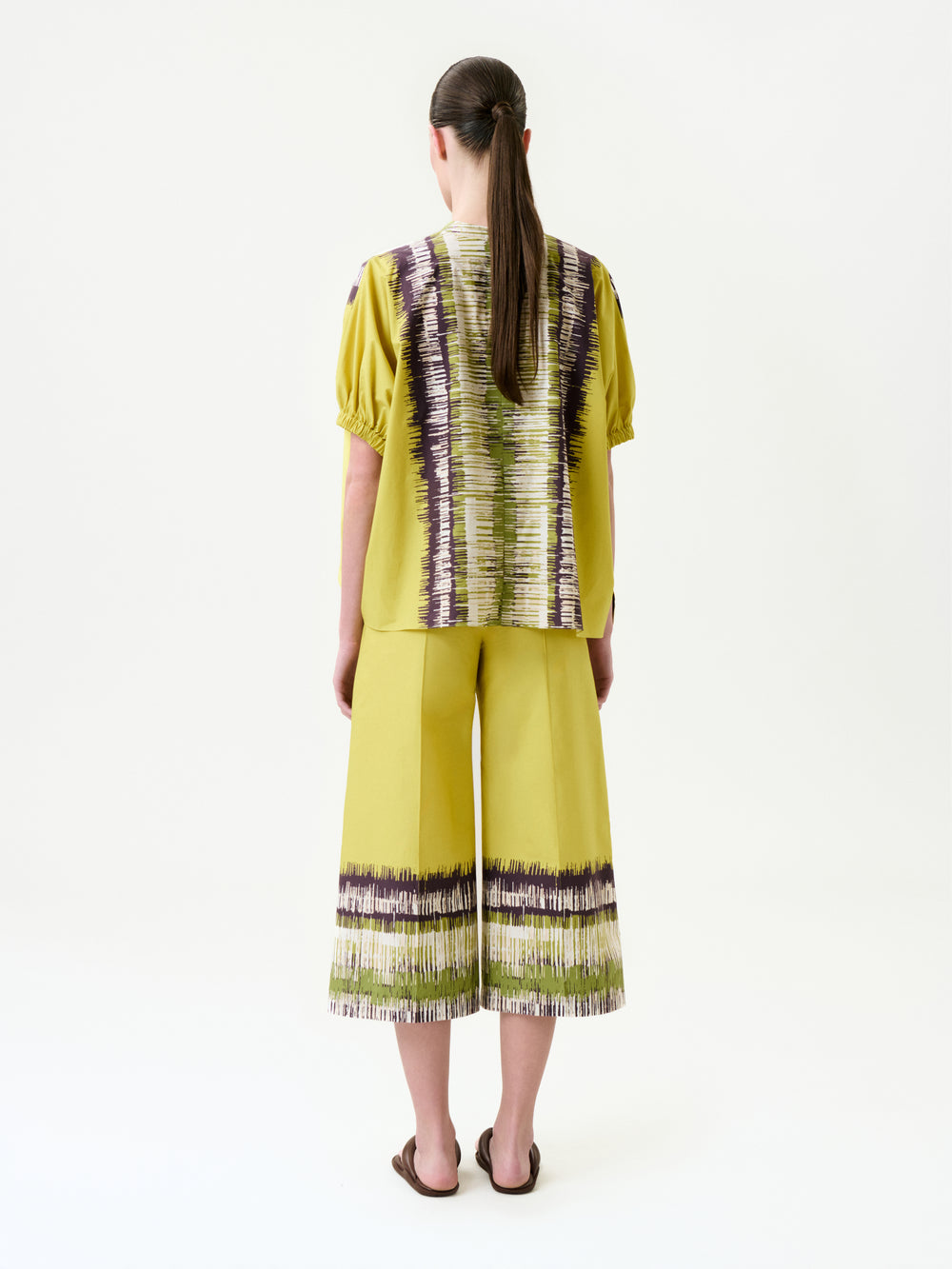 A person with long brown hair, wearing a yellow short-sleeved top and the Ilma Pant—wide-leg pants with purple and white abstract stripes in Italian fabric—stands facing away against a plain white background.