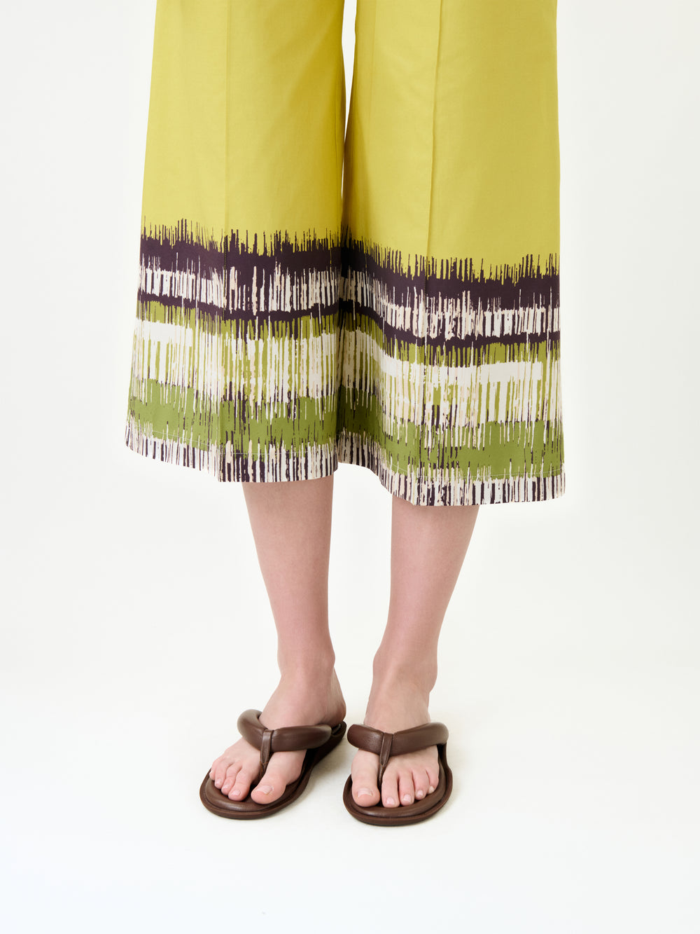 A person models the Ilma Pant—wide-leg yellow-green pants in Italian fabric with a brushstroke hem pattern—on a white background, highlighting their brown sandals.