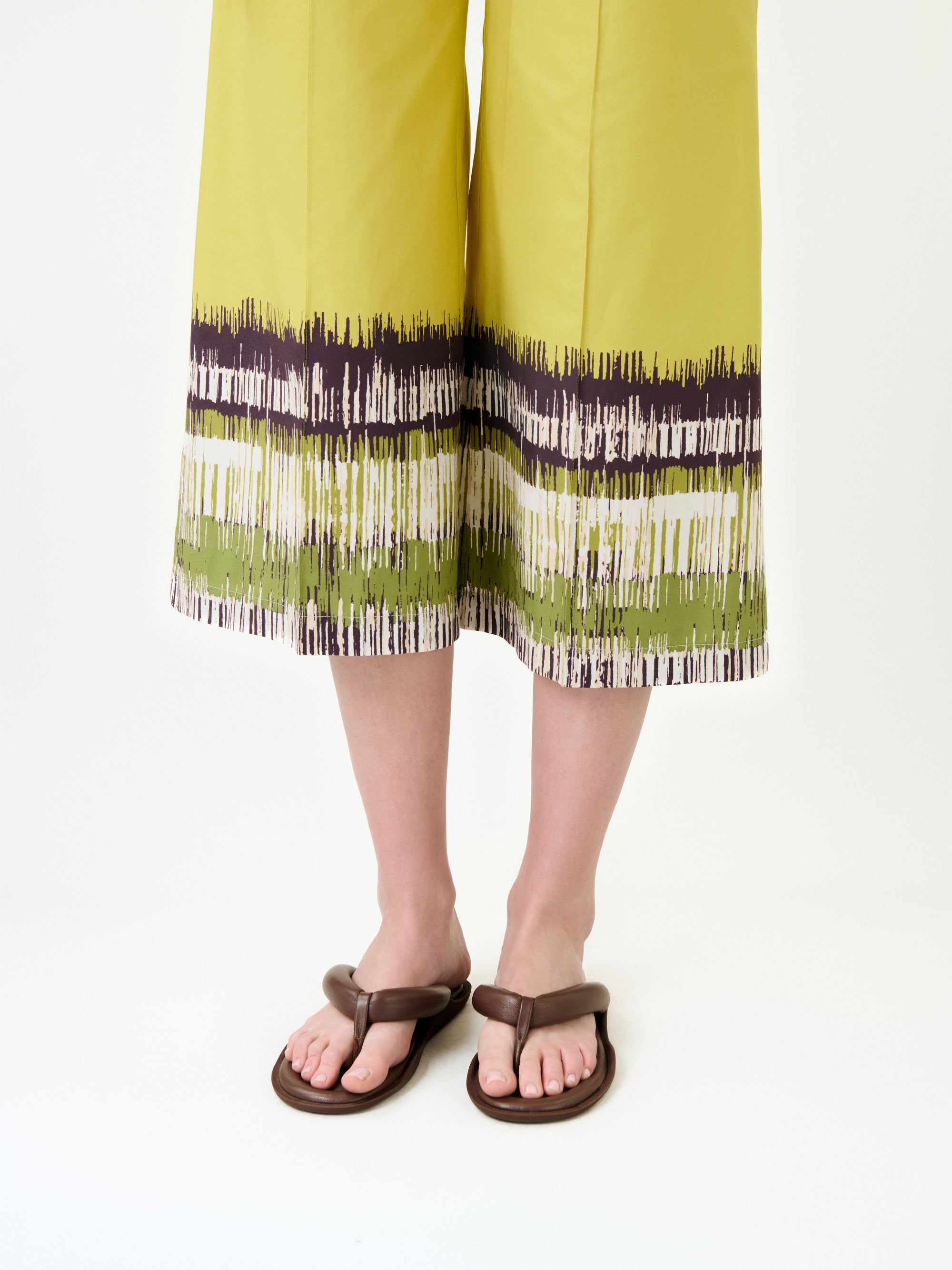 A person models the Ilma Pant—wide-leg yellow-green pants in Italian fabric with a brushstroke hem pattern—on a white background, highlighting their brown sandals.
