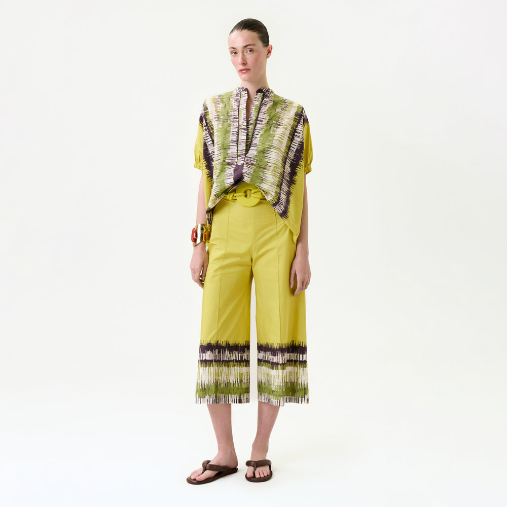 A woman stands against a white background wearing the Ilma Pant, wide-leg cropped pants with patterned hems, paired with a matching short-sleeve patterned top. Her hair is tied back and she completes the look with sandals.