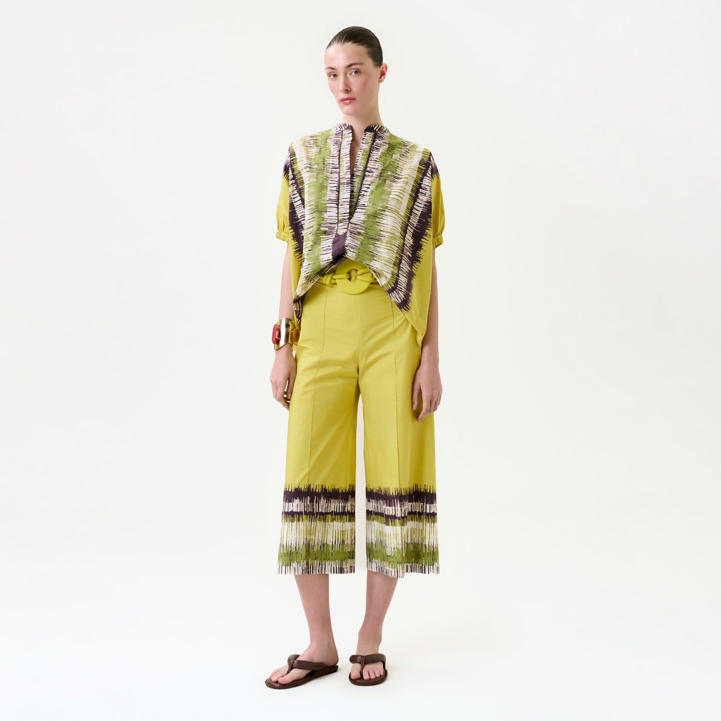 A woman stands against a white background wearing the Ilma Pant, wide-leg cropped pants with patterned hems, paired with a matching short-sleeve patterned top. Her hair is tied back and she completes the look with sandals.