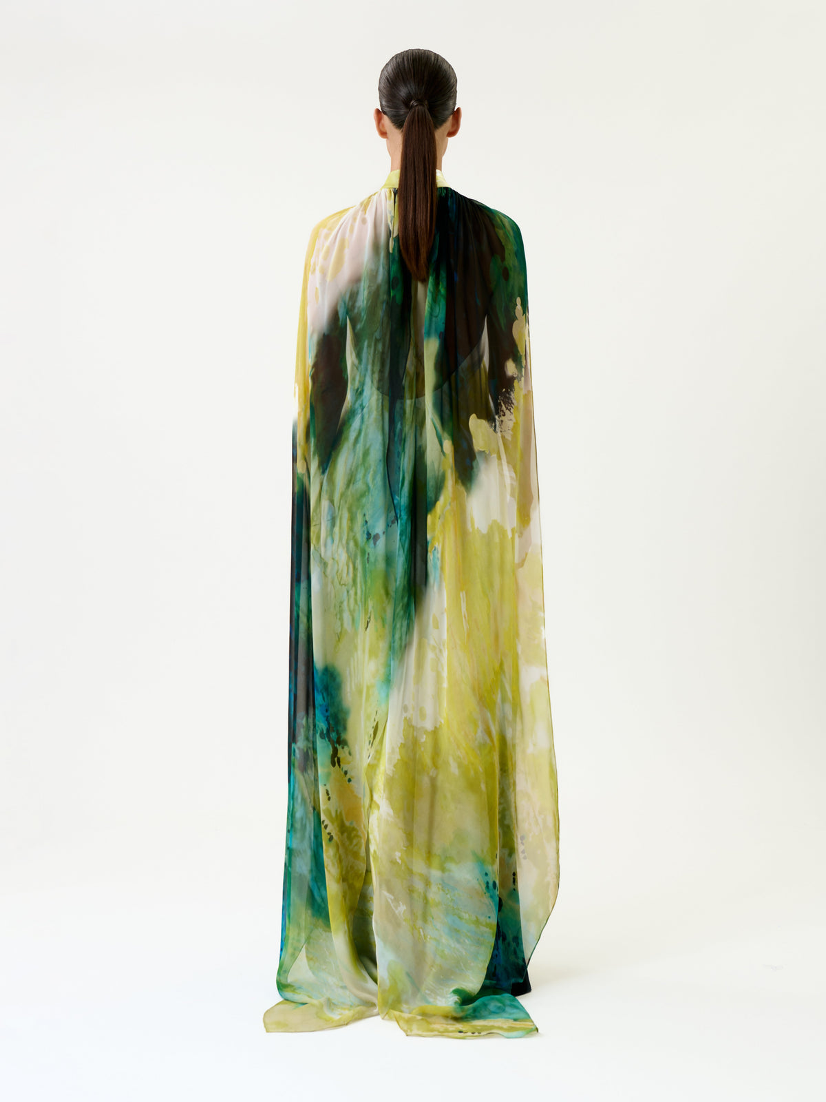 A person with brown hair in a low ponytail is wearing the Sabina Dress—a long, flowing gown with yellow and green patterns—standing against a plain white background, facing away from the camera.