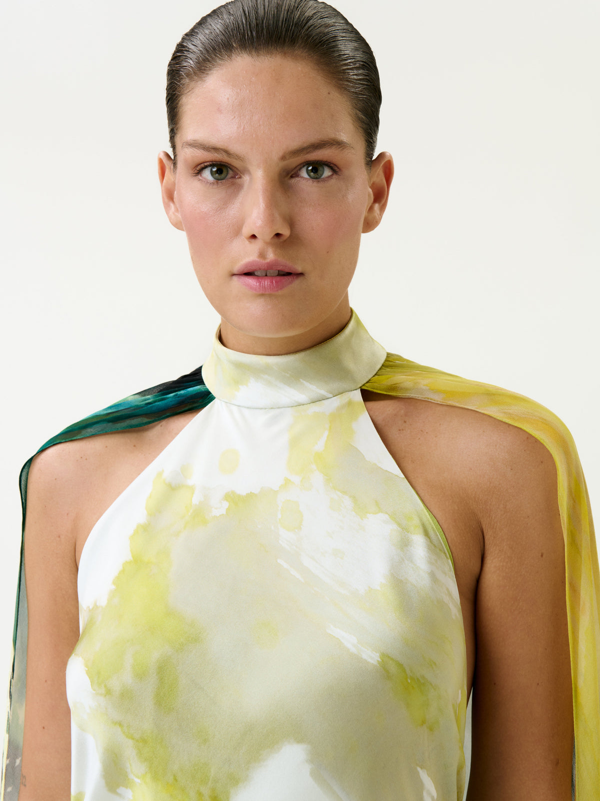 A woman with slicked-back hair wears the Sabina Dress, a sleeveless, high-neck style featuring yellow and green watercolor patterns, against a plain background.