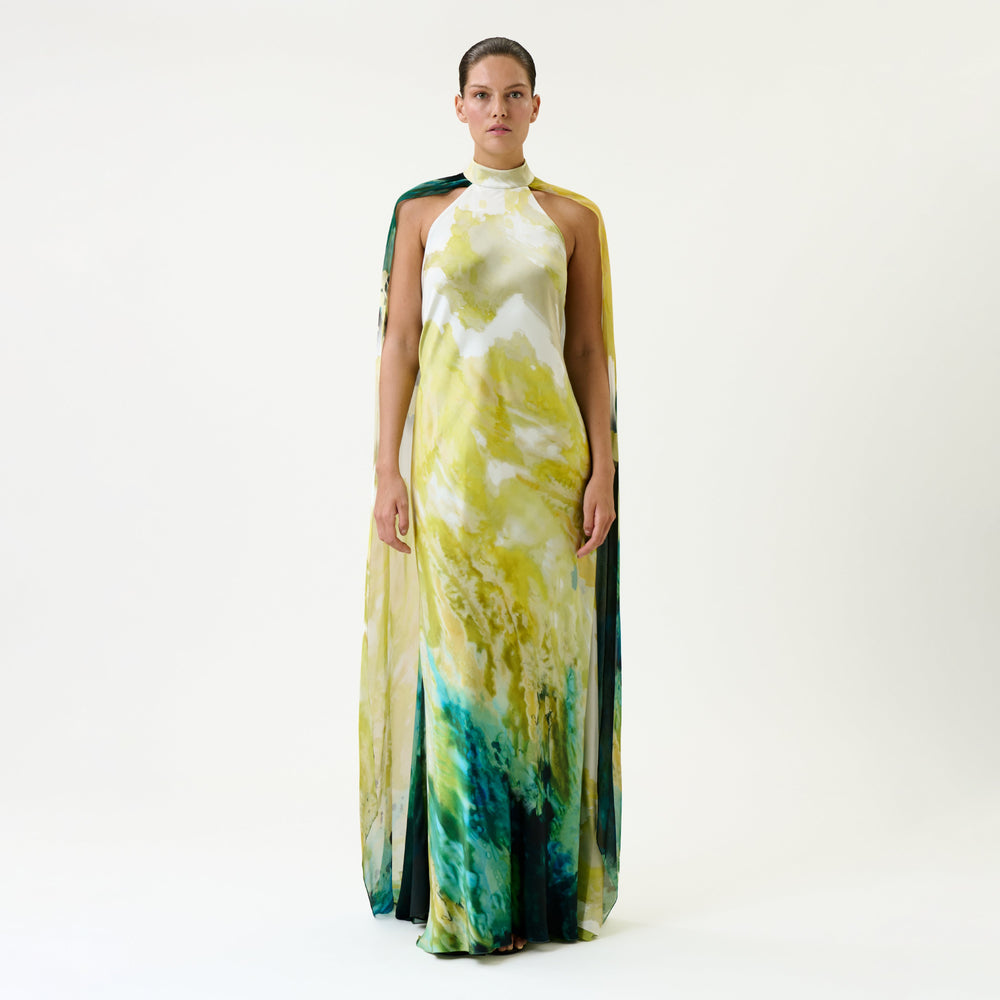 A person stands against a plain white background wearing the Sabina Dress, a long, sleeveless gown featuring a yellow, white, and green marble pattern with a matching cape.