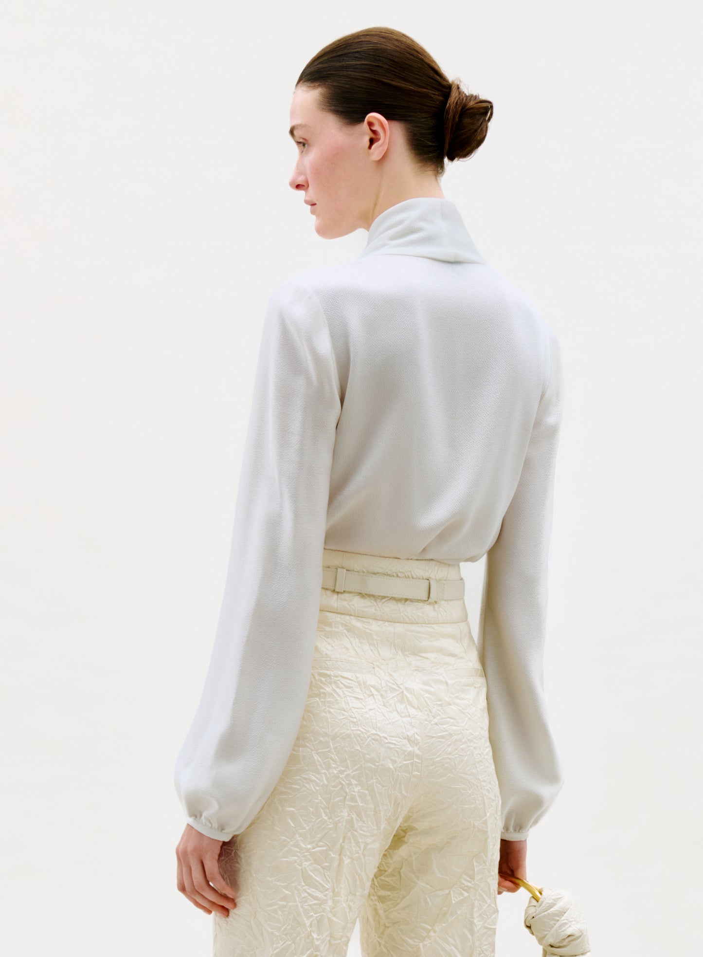 A woman with brown hair in a low bun models the Tea Blouse, a long-sleeve white top, paired with high-waisted cream pants; she stands at an angle against a plain light background, holding a small light item in her left hand.