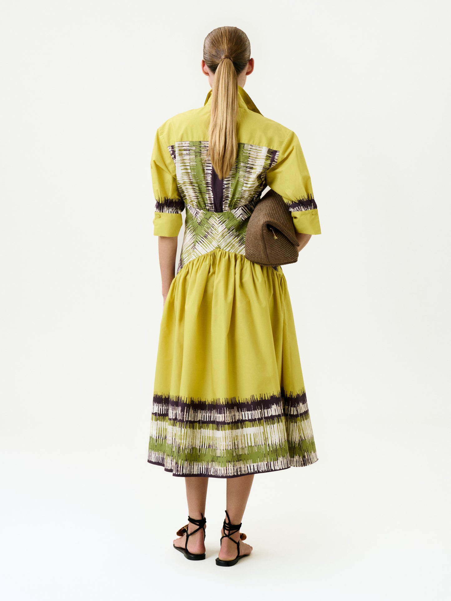 A woman with long, straight hair in a low ponytail is seen from behind against a plain white background, wearing the yellow Indy Dress crafted from Italian fabric with patterned accents, black sandals, and holding a brown textured clutch.