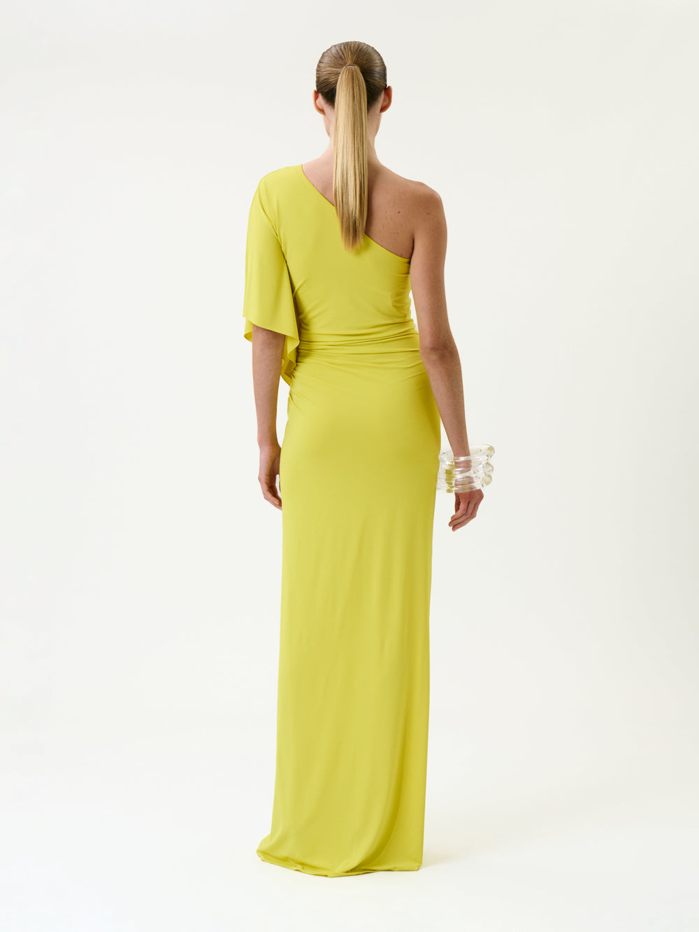 A person with a ponytail wears the Doli Dress—a bright yellow, one-shoulder, floor-length gown—paired with a chunky clear bracelet, standing against a plain white background and facing away from the camera.