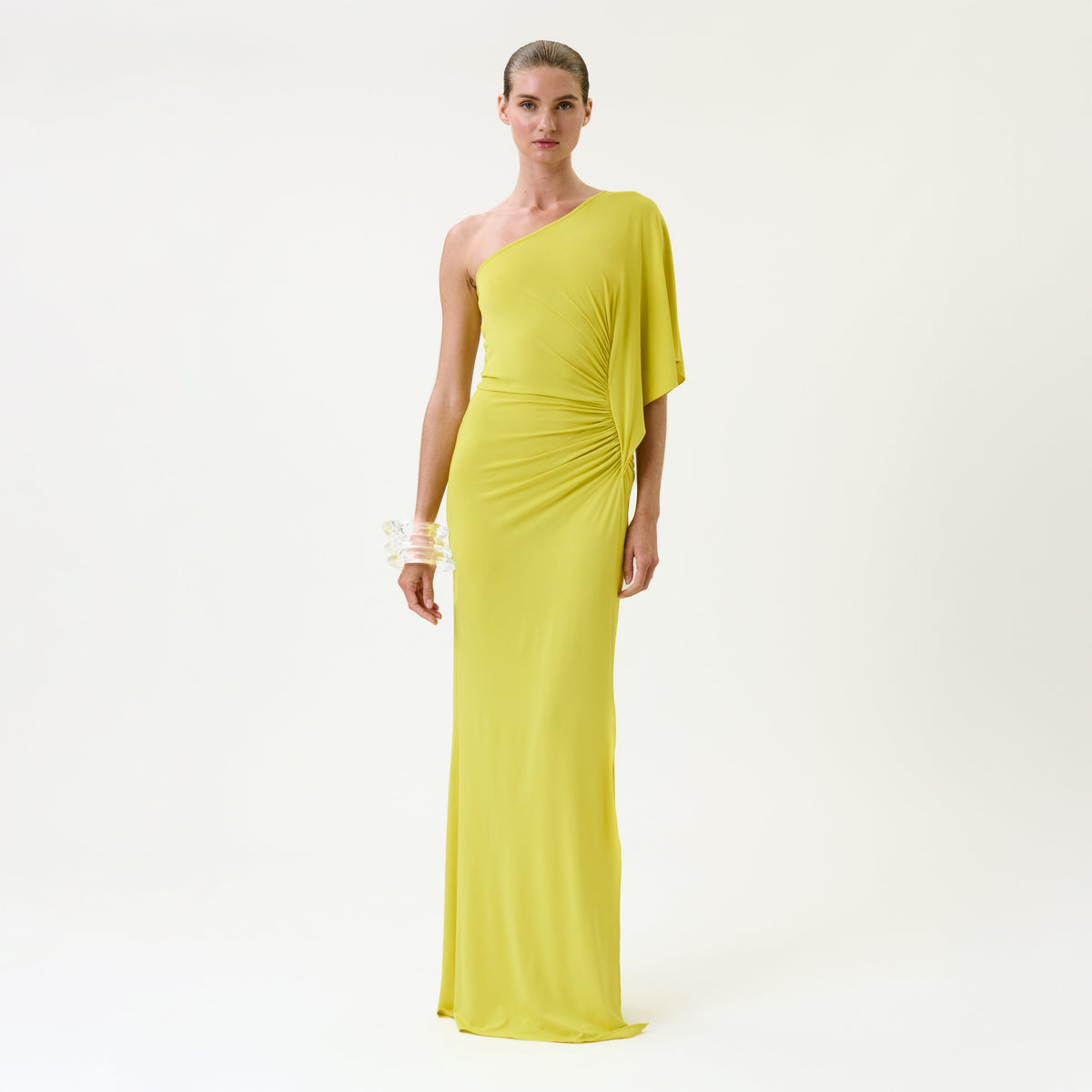 A woman stands against a plain white background wearing the Doli Dress, a vibrant yellow, one-shoulder, floor-length gown. Her hair is slicked back and she accessorizes with a bracelet on her left wrist.