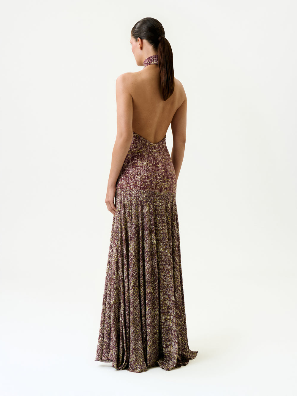 A woman with dark hair in a ponytail faces away from the camera, wearing the Leonore Dress—a halter-neck, backless maxi dress featuring a multicolored textured pattern, made in Colombia—against a plain white background.