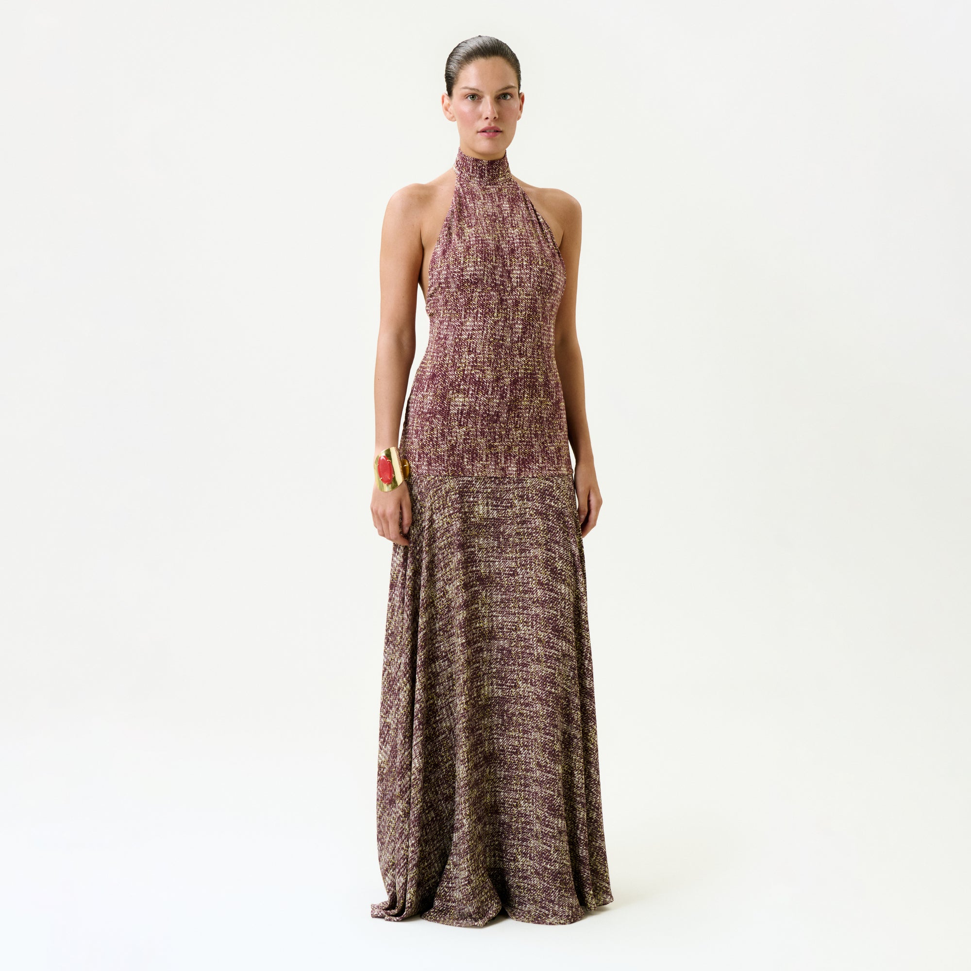 A woman with her hair pulled back models the Leonore Dress—a sleeveless, halter-neck, floor-length maroon and beige textured maxi made in Colombia—against a plain white background, accessorized with a chunky bracelet on her left wrist.