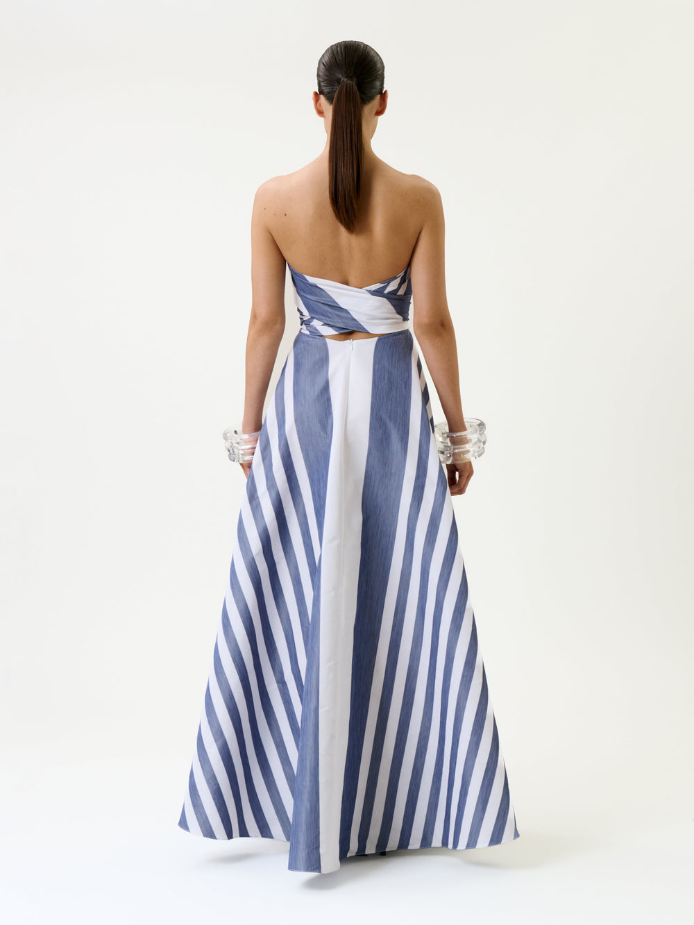 A woman stands with her back to the camera, wearing the Leyna Dress—a strapless blue and white striped gown featuring a fitted bodice and flared skirt. Her hair is styled in a ponytail, and she accessorizes with large clear bangles on both wrists.