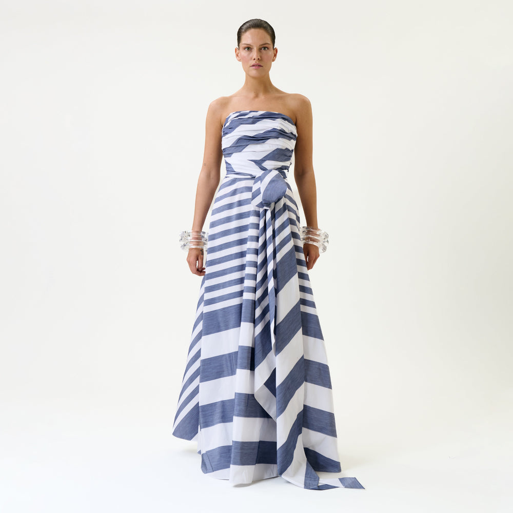 A woman stands against a plain background wearing the Leyna Dress, a strapless, full-length design featuring bold blue and white diagonal stripes, paired with clear chunky bracelets on both wrists.