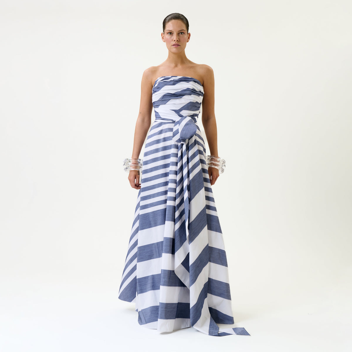 A woman stands against a plain background wearing the Leyna Dress, a strapless, full-length design featuring bold blue and white diagonal stripes, paired with clear chunky bracelets on both wrists.