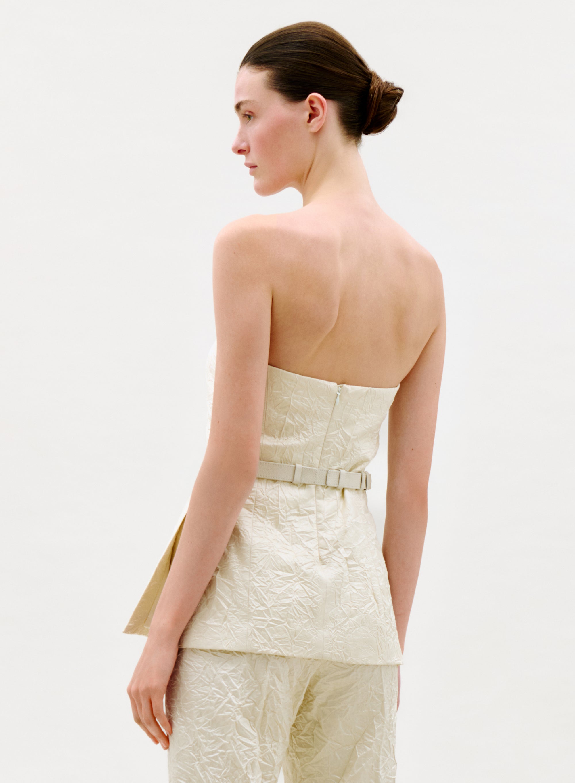 A woman with fair skin and brown hair in a low bun is seen from the back and side wearing the cream-colored, textured Astor Top with matching pants against a plain white background.