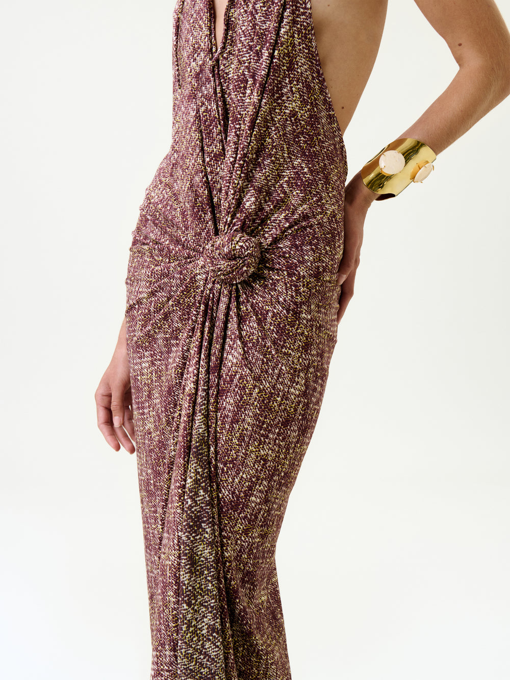 A person wears the Abril Dress, a sleeveless maroon and gold maxi with textured fabric and a knotted waist. Their right hand, adorned with a chunky gold bracelet, rests on their hip against a plain white background.