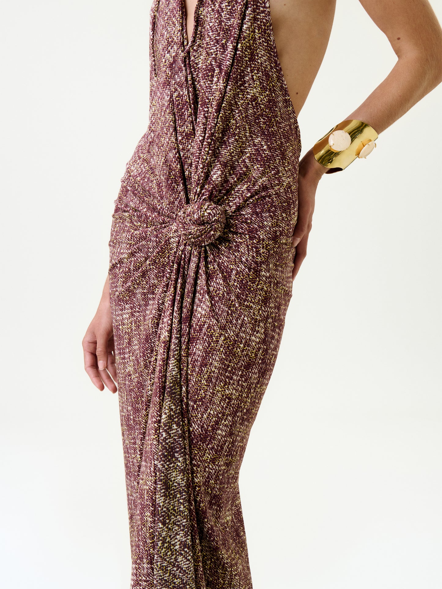 A person wears the Abril Dress, a sleeveless maroon and gold maxi with textured fabric and a knotted waist. Their right hand, adorned with a chunky gold bracelet, rests on their hip against a plain white background.