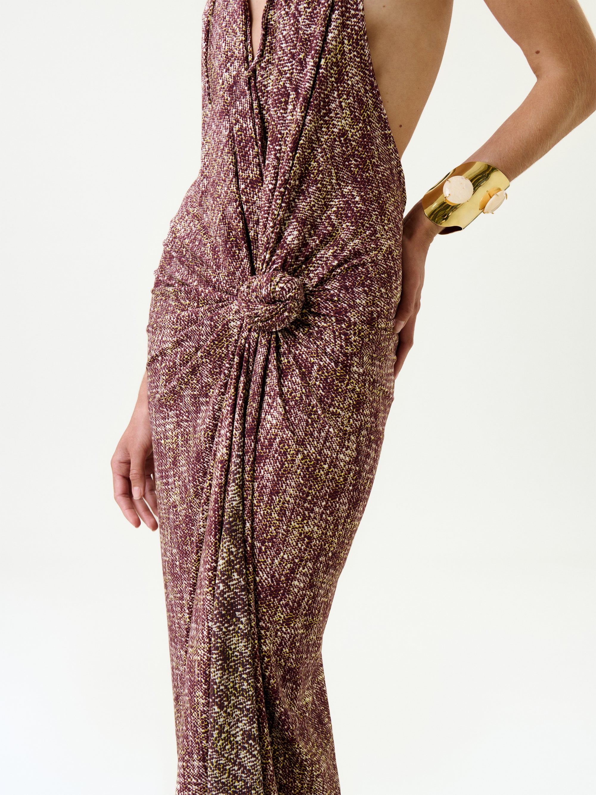 A person wears the Abril Dress, a sleeveless maroon and gold maxi with textured fabric and a knotted waist. Their right hand, adorned with a chunky gold bracelet, rests on their hip against a plain white background.