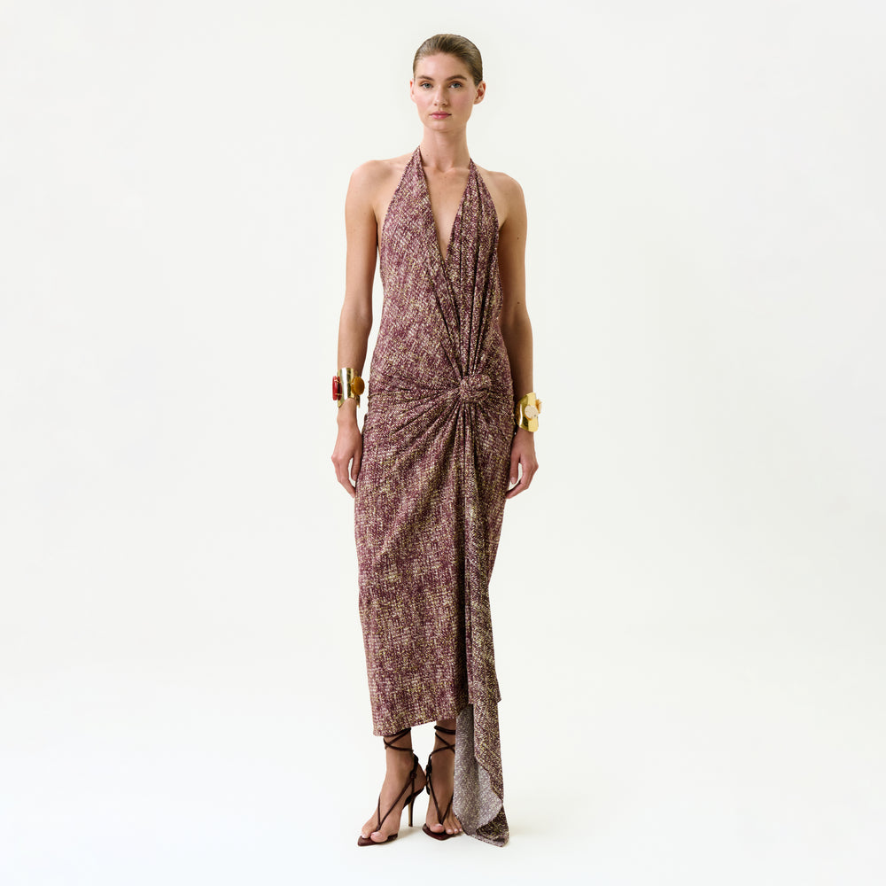 A woman stands against a plain background wearing the Abril Dress, a sleeveless halter-neck brown and purple patterned maxi dress from Colombia with a knot detail at the waist. She pairs it with minimal accessories and strappy high-heeled sandals.