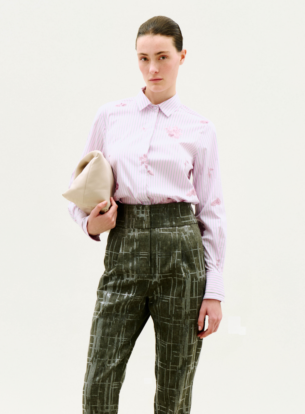 A woman with dark hair pulled back and a neutral expression stands against a plain background, wearing the Linette Blouse, a light pink striped button-up shirt, paired with high-waisted dark green patterned trousers and holding a beige clutch.