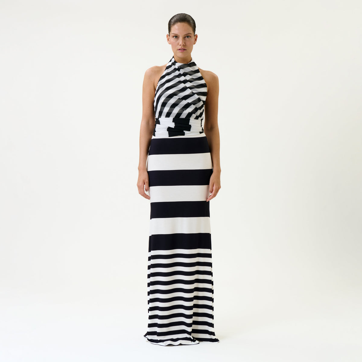 A woman stands facing forward wearing the Tove Dress, a long, sleeveless, black-and-white striped dress with a halter neckline. The background is plain and white.