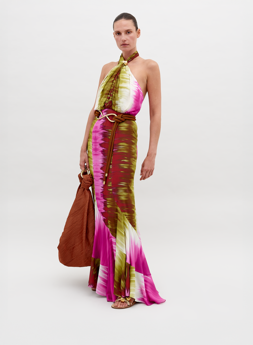 A person dressed in a Raisa Dress Violeta Lime Tie Dye with pink, green, and white patterns, holding a large brown bag, posing against a plain background. Pre-order by November 15th 2024 to ensure the perfect ship-by date.