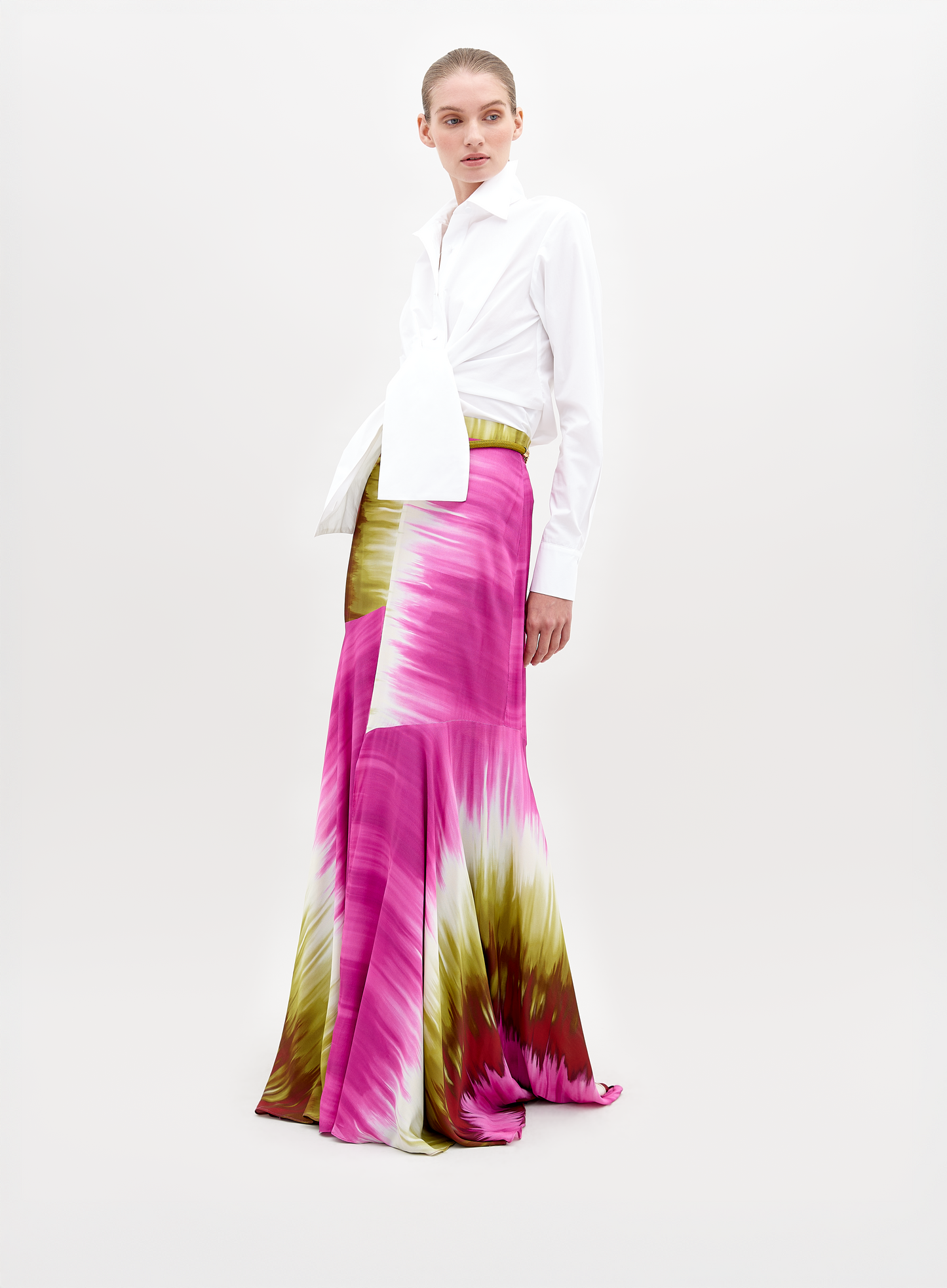 A person stands wearing a white collared shirt tucked into a long, flowing Shane Skirt Violeta Lime Tie Dye with patterns in pink, green, and white. The background is plain and light-colored. Secure this stylish look by placing a pre-order now to ensure it arrives before the ship by date of November 15th, 2024.