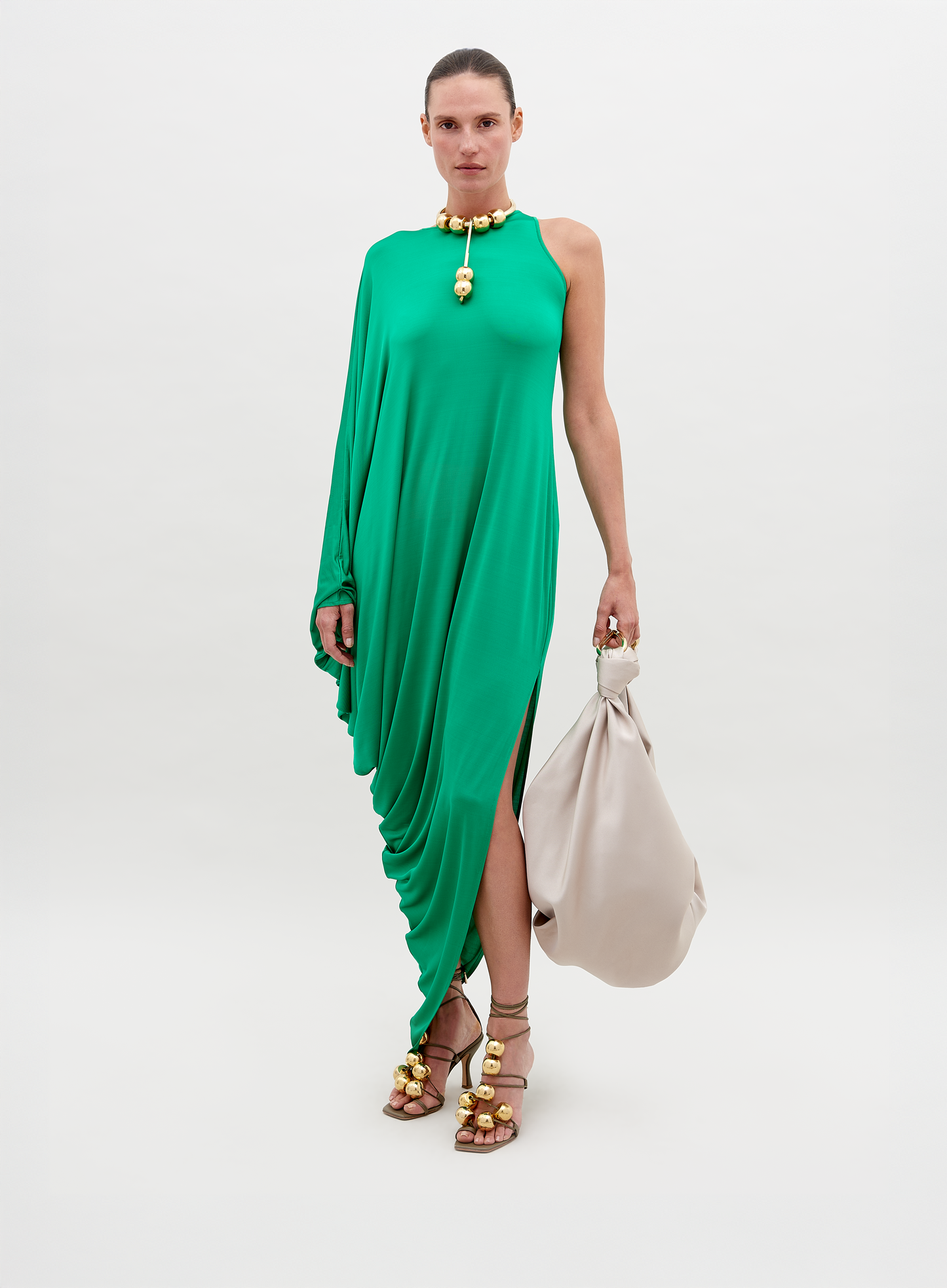 A person stands against a plain background, wearing a Golda Dress Green with gold accessories, holding a large beige handbag. Pre-order now for shipping by November 15th, 2024.