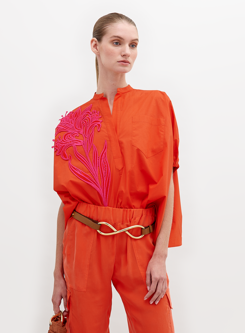 A woman stands against a white background, wearing an orange outfit with a pink floral pattern on the upper left side and a gold belt. The Susanne Blouse Scarlet is available for pre-order until November 15th, 2024.