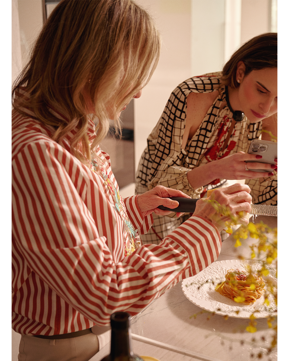 Two women in a kitchen, one taking a photo of food on a plate.