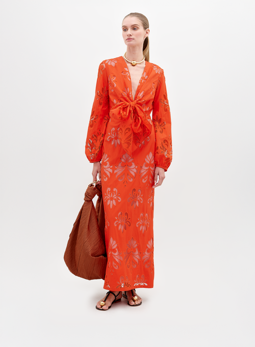 A person stands in a long, bright orange Battia Dress Rouge Embroidery with floral patterns and a front bow tie. They carry a large, rust-colored bag and wear gold jewelry and black sandals. To ensure you receive this stunning ensemble by the desired ship-by date, consider placing your pre-order for 2024 today.