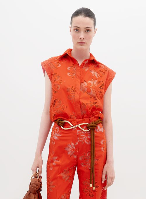 A person stands wearing a vibrant orange sleeveless top and matching pants with floral patterns, accessorized with a brown belt and holding a brown handbag. Pre-order the Rina Blouse Rouge Embroidery now to ensure it ships by the date of November 15th, 2024.