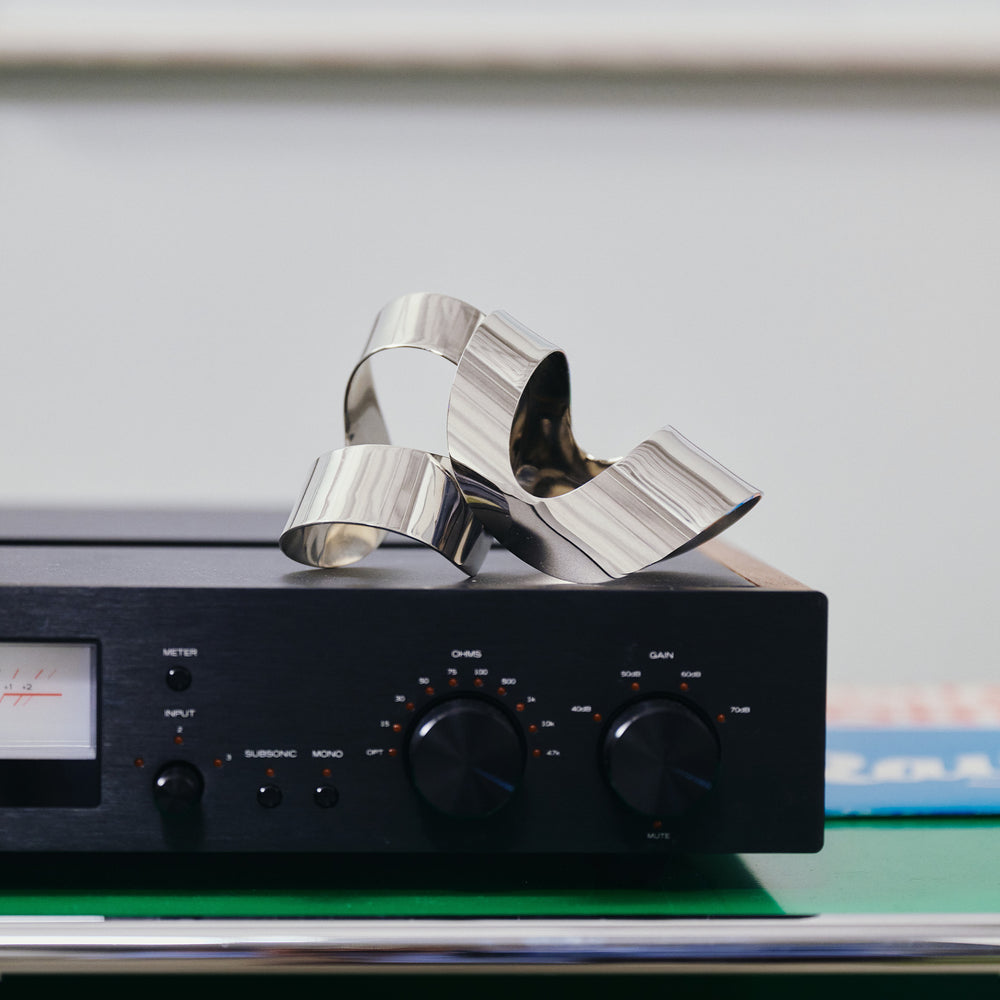 A polished silver metallic sculpture with flowing, ribbon-like curves resting on top of a black audio amplifier or receiver, photographed in a clean indoor setting.