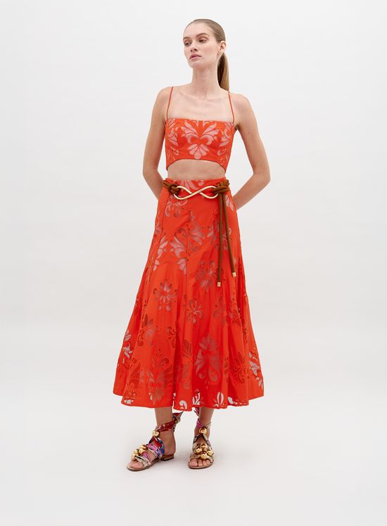 A woman stands wearing a Grettel Skirt Rouge Embroidery with a bright orange floral-patterned two-piece outfit and colorful sandals. She poses against a plain white background, available for pre-order starting November 15th for the 2024 collection.