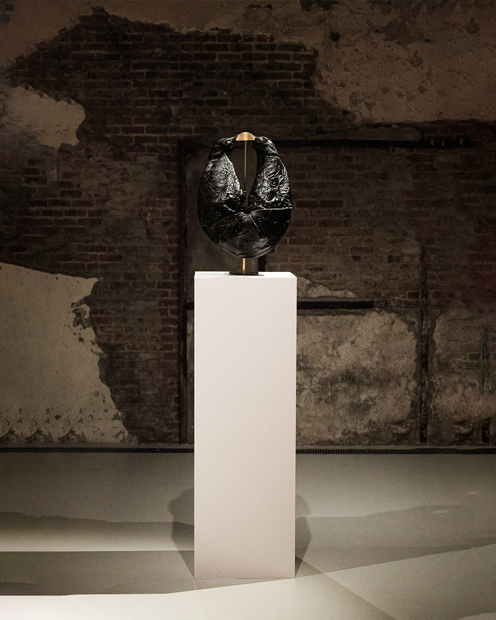 A black sculptural art piece displayed on a tall white pedestal in front of a rustic brick and plaster wall.