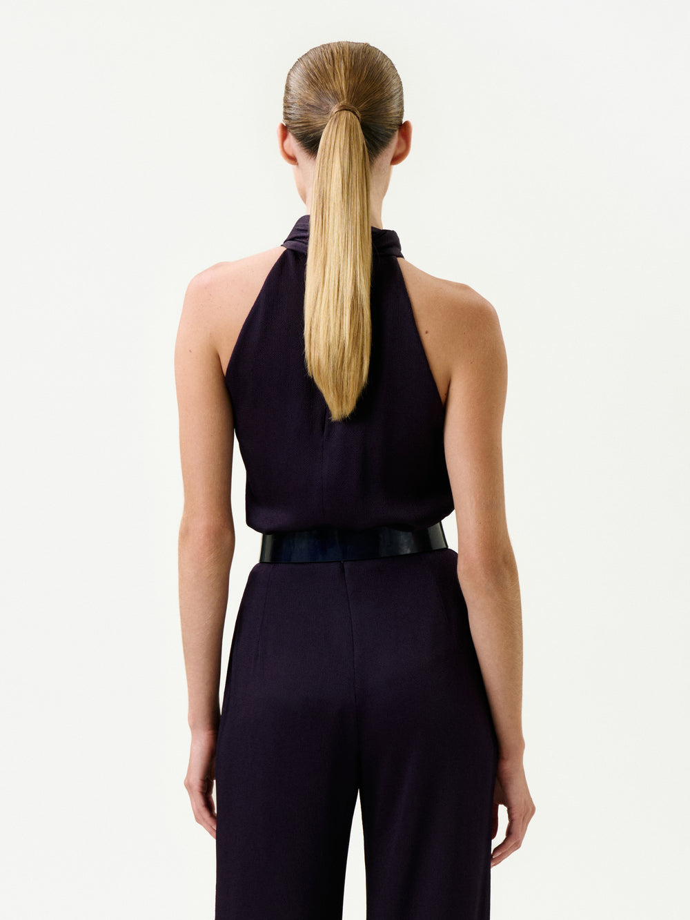 A woman with straight, long blonde hair in a low ponytail is shown from the back, wearing the sleeveless Naili Blouse with a high collar and black belt, crafted from luxurious Italian fabric. She stands against a plain, light background.
