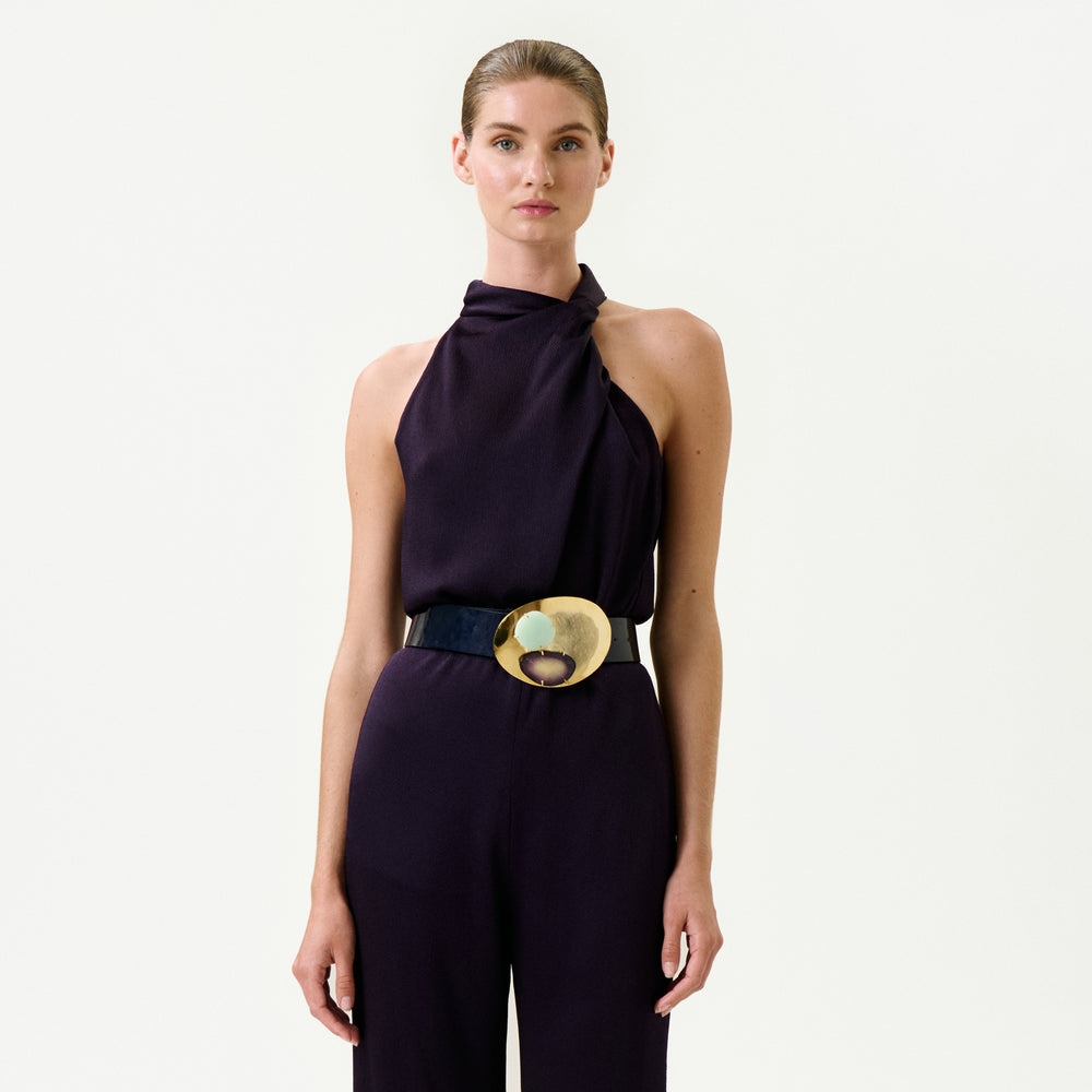 A woman stands against a white background wearing the Naili Blouse—a sleeveless, dark purple Italian-fabric top with a high neckline—paired with a wide black belt with a large, round gold buckle. Her hair is pulled back and she has a neutral expression.